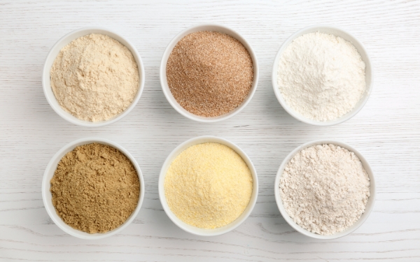 Six small white bowls, filled with different types of flour, on a light white wood background